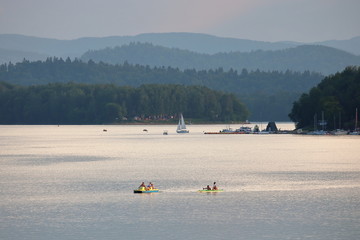 Jezioro Solińskie, Polska, lato, na wodzie jachty, rowerki wodne, kajaki, na drugim brzegu zielone wzgórza, tafla wody lśni srebrzyście