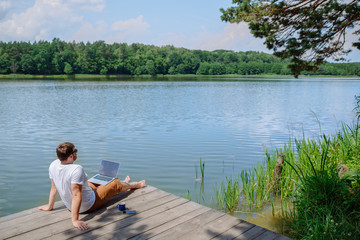 man working on laptop while sitting on wooden dock. legs in river water