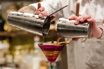 Professional bartender pouring cocktail from the shaker into the glass. Barman holding in hands cocktail tool. Mixing alcoholic drink process. Close up image with part of human body. 