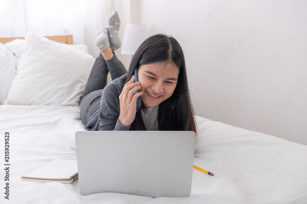 Poster Asian woman laying on bed talking on mobile phone while browsing data on laptop in bedroom.work at home.e-larning concept.study online.
