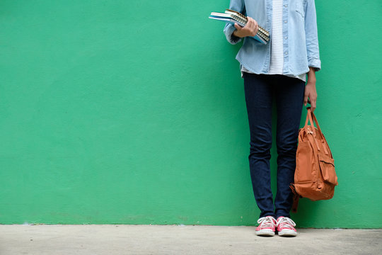 Student girl holding books and school bag standing over green wall background with copy space, education, back to school concept