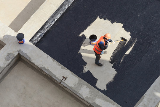 The worker in overalls applies an insulation coating on the concrete surface. View from above.