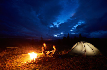 Fototapeta na wymiar Happy couple man and woman travellers having a rest at night camping in the mountains, sitting near burning campfire and illuminated tourist tent under beautiful evening cloudy sky. Tourism concept