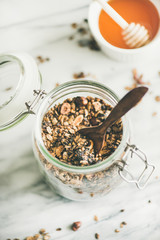 Fototapeta premium Healthy breakfast preparation. Buckwheat and chocolate granola with hazelnuts in glass jar over light marble background, top view. Clean eating, vegan, vegetarian food concept