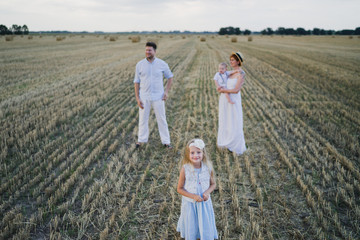 Happy stylish caucasian family, mother, father, daughter and son, walking together in summer wheat field. Mother and father playing with children outdoors. Love, parenthood, childhood, happiness.