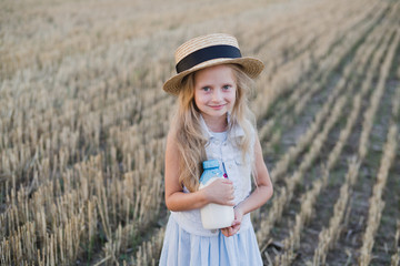 Happy cute cheerful caucasian little girl wearing hat and walking in summer wheat field holding a bottle of milk. Beautiful girl portrait outdoors. Childhood, beauty, happiness concept.