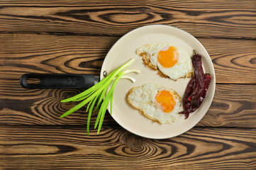 Two fried eggs in metal beige pan and green fresh onion and dried red hot peppers on old wooden brown table. Top view