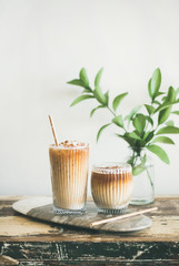 Iced coffee in tall glasses with milk and straws on board, white wall and green plant branches at...