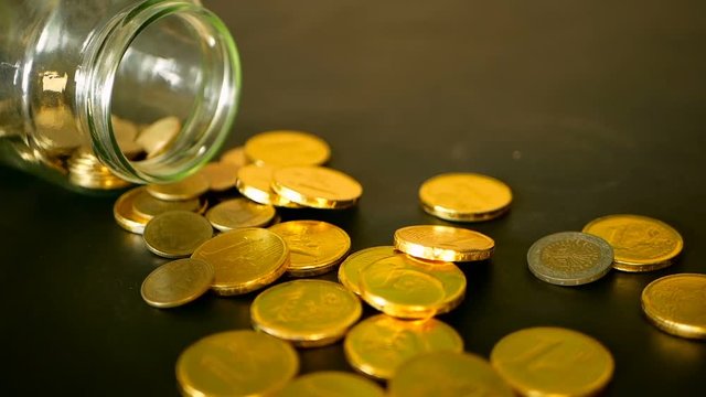 Yellow coins fell out from jar. Symbol of investing, keeping money concept. Collecting cash conis in glass tin as moneybox. Close-up still life with gold coins on black table and rotating penny.