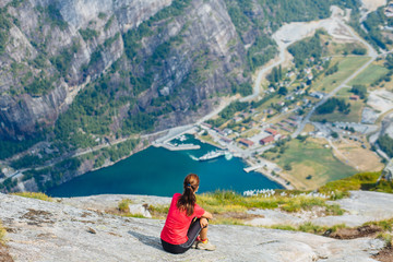 Adventure, travel, tourism, hike and people concept - cheering woman in pink t-shirt sitting over mountains nature background. Female admiring views, relaxing during climbing on Kjeragbolten, Norway