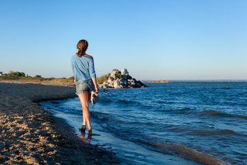 A cheerful dark-haired woman in jeans and a striped T-shirt smiles, walks along the beach and enjoys the bright sun on a summer day. Concept of summer holidays at sea and live style