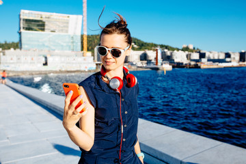 Obraz premium Young hipster business woman traveler walking in Oslo city on quay, listening to music on red headphones, street style, urban background, holding smart phone, happy, smiling, taking selfie photo.
