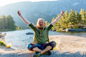 Emotional portrait of handsome beard blond long haired man traveler holding hands up over beautiful scandinawian landscape backround in sunset. Adventire and freedom concept.