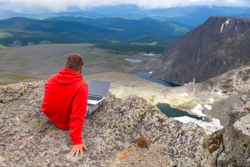Naklejka premium Young tourist man working on a laptop and sitting on the top of the mounting and looking at a beautiful landscape. 