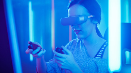 Pro Gamer Girl Wearing Virtual Reality Headset Plays Online Video Game with Joysticks / Controllers. Cool Blue Neon Colors in the Room.