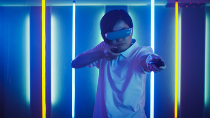 East Asian Pro Gamer Wearing Virtual Reality Headset Plays Online Video Game Shooter using Joysticks / Controllers as Shotgun / Gun. Cool Retro Neon Colors in the Room.
