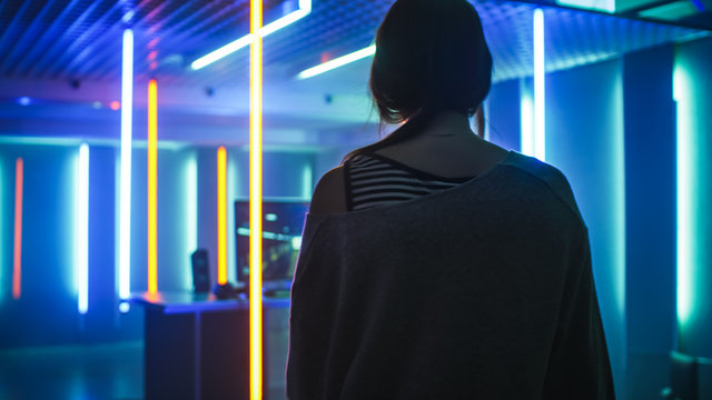 Back View Shot of the Beautiful Professional Gamer Girl Looking at Her Room in Retro Neon Lights with Her Personal Computer. Casual Cute Geek wearing Glasses.