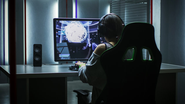 Beautiful Professional Gamer Girl Playing in First-Person Shooter Online Video Game on Her Personal Computer. Casual Cute Geek Girl Wearing Headset. Dark Room in Gray Tone without Neon Lights.