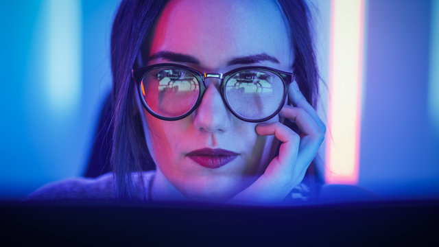 Portrait of the Beautiful Young Girl Sitting Before Computer Screen, Browsing in Internet, Playing Online Games, Streaming. Cute Girls Wearing Glasses in the Cool Retro Neon Lit Room.