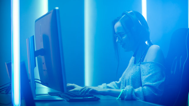 Beautiful Professional Gamer Girl Plays in First Person Shooter Video Game on Her Personal Computer, Casual Cute Geek wearing Glasses, talks with Her Teammates. Neon Lights Room. eSport Cyber Games