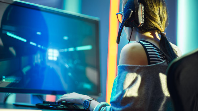 Shot of the Beautiful Pro Gamer Girl Playing in FPS Video Game on Her Personal Computer, Casual Cute Geek wearing Glasses and Headset. Neon Room. Playing Online Games.