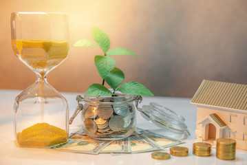 Real estate investment or property ladder. Home mortgage loan rate. Saving money for future concept. Plant growing out of coins in glass jar with dollar banknotes, hourglass and house model on table