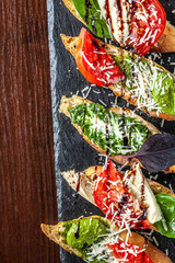 Italian bruschettes with mozzarella cheese, tomatoes, pesto sauce and parmesan cheese, on a black stone, on a wooden table. Copy space, selective focus