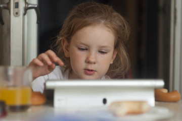 The little girl combines breakfast and games on a tablet. Modern generation.