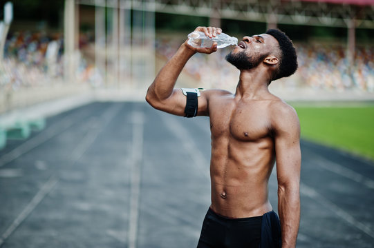 African american male athlete sexy sport bare torso man with running sports arm case for mobile phone, posed at stadium and drinking water from bottle.