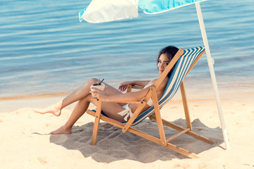 attractive girl in bikini with coconut cocktail resting on beach under umbrella