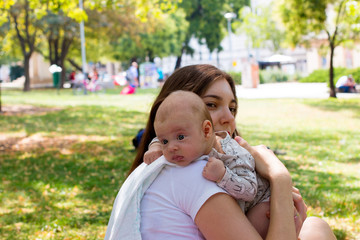 Portrait of beautiful baby resting her head on the mother arm, young mom is caring her infant in burping position after breastfeeding, outdoor city park scenery around