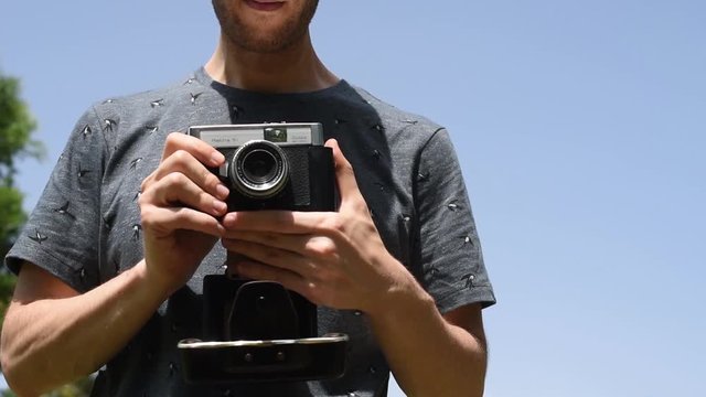 Detalle de fotógrafo con camiseta de pájaros haciendo fotos con una cámara antigua de película. El fotógrafo se encuentra al aire libre en medio del campo bajo un cielo azul despejado.