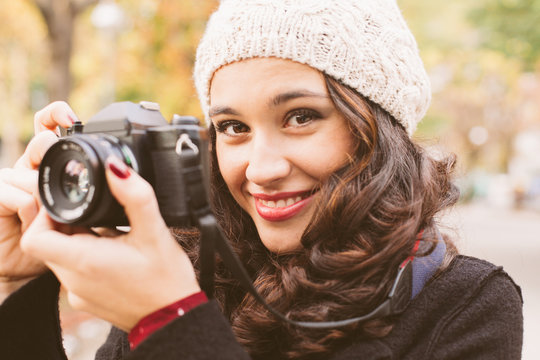 Young beautiful woman with a woolen cap taking pictures with a retro camera in the city in autumn