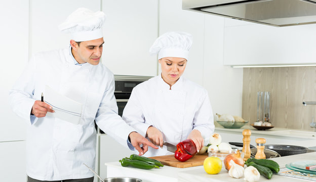 Female and male young cooks with paper recipe in uniform working