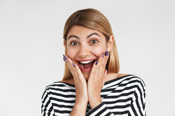 Fototapeta premium Studio shot of funny emotional young Caucasian woman in striped top exclaiming Yes to unexpected marriage proposal. Excited girl holding hands on her cheeks, celebrating success or victory