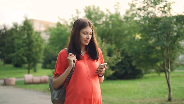 Beautiful pregnant girl expecting mother is walking in city park and using smartphone, young woman is holding device and touching screen, summer nature is visible.