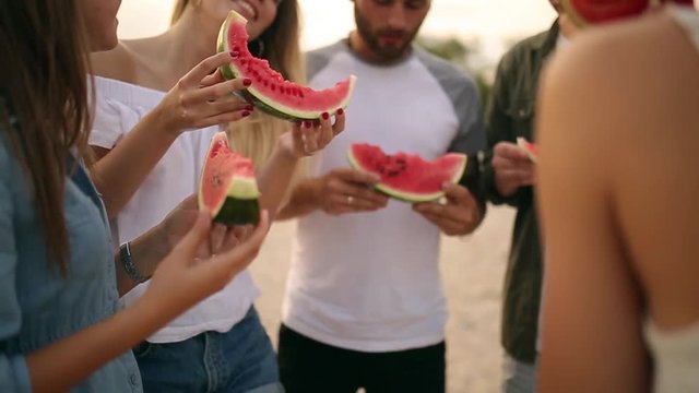 Happy Friends Eating Watermelon Standing on Sandy Beach and Chatting. Young Men and Women Wearing Blue Jeans Shorts Near Sea on Vacation. Friendship and Summer Concept