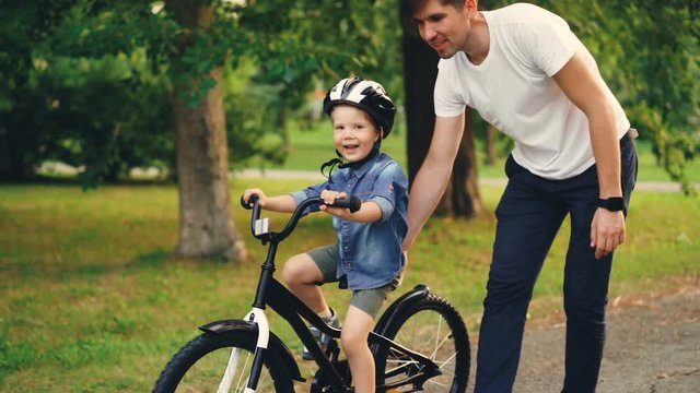 Slow motion of cheerful guy caring father teaching his small son to ride bicycle in park. Cute boy is cycling while young man is holding bike and helping child.
