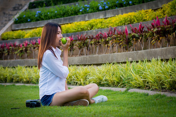 Young woman wear white clothing and holding an apple sitting on the grass.