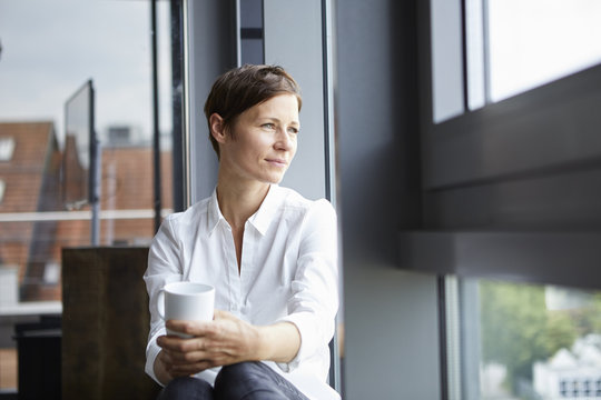 Businesswoman sitting in office with cup of coffee looking out of window