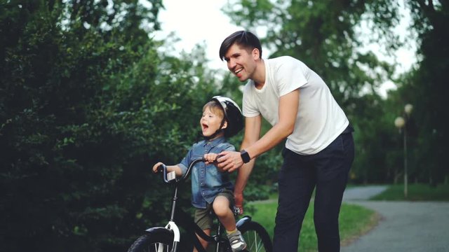 Slow motion of happy young man loving father teaching his child to cycle in green park in summer, little boy is laughing, shouting and enjoying weekend with dad.