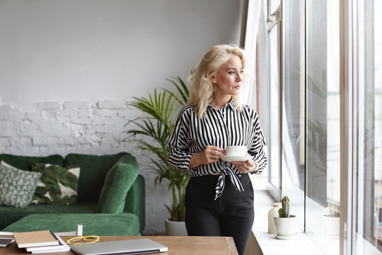 Serious Fifty Year Old Female Freelancer In Striped Shirt And Black Trousers Having Coffee Break, Standing At Her Workplace With Cup, Looking Through Window, Using Portable Computer For Remote Work