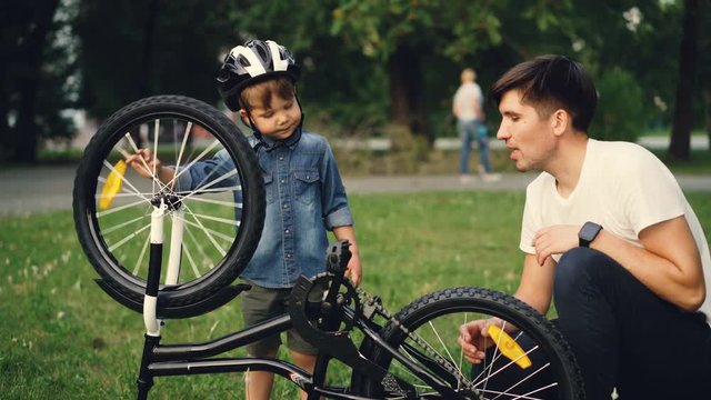 Little boy is spinning bicycle wheel and pedals while his father is talking to him on lawn in park on summer day. Family, leisure and active lifestyle concept.