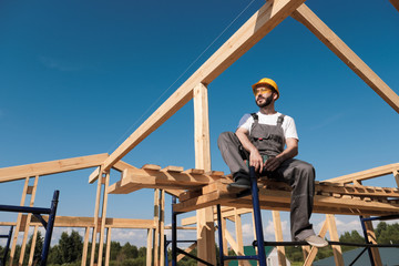 The man builder sits on the edge of the roof of the frame house, in a yellow helmet and gray...