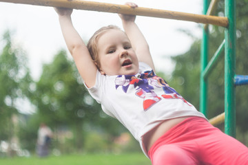 Obraz premium Girl Hanging and swinging on hands, on a crossbar on a children's playground. The girl is looking directly at the camera.