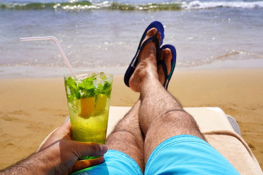 Man relaxing on a sunbed and sunbathing with a cold mojito drink on the beach sand. Summer vacation concept image.