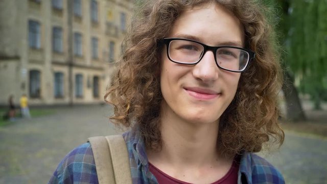 Young handsome man in glasses with curly hair smiling at camera and standing on street near college building, happy