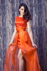 Young brunette woman in stylish orange silk dress posing on wooden background