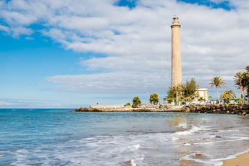 Tenerife, Canarias islands/ Spain - July 22, 2018: View of empty beach of tourists and high lighthouse on Canarias, view of bay with palms. Blue Atlantic ocean and beach on Gran Canaria. - obrazy, fototapety, plakaty