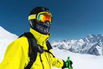 Close-up portrait of a skier in a mask and helmet with a closed face against a background of snow-capped mountains and blue sky